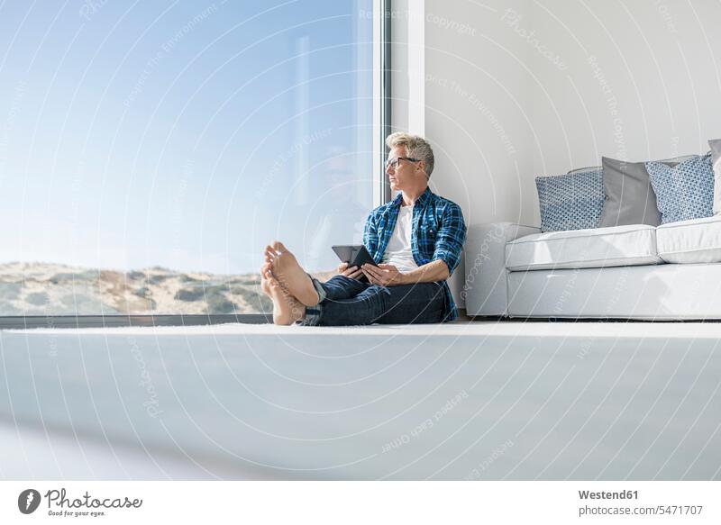 Casual businessman sitting in front of window in modern home holding tablet business life business world business person businesspeople Business man
