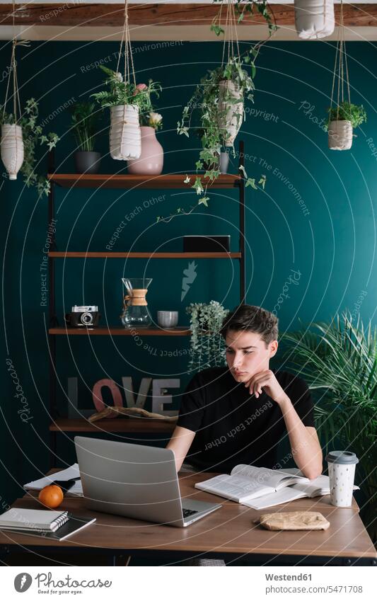 Handsome young man using laptop while doing homework at table against rack color image colour image indoors indoor shot indoor shots interior interior view