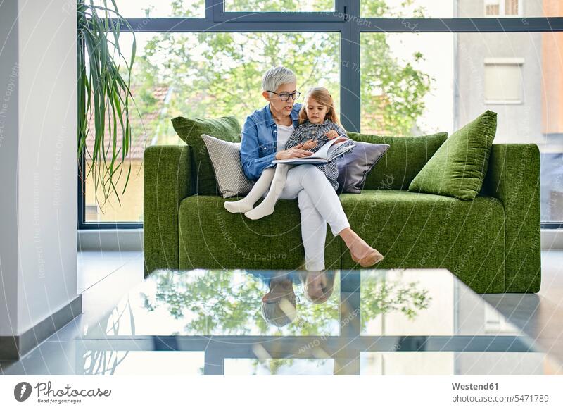 Grandmother sitting on couch with granddaughter, reading book together generation books couches settee settees sofa sofas Seated Secure at home indoor