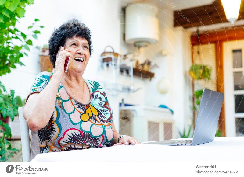Cheerful senior woman talking over smart phone while sitting by laptop in yard color image colour image Spain leisure activity leisure activities free time