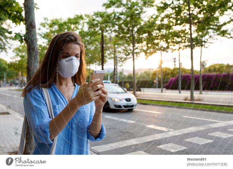 Young woman wearing mask using smart phone while standing on street in city color image colour image Spain casual clothing casual wear leisure wear