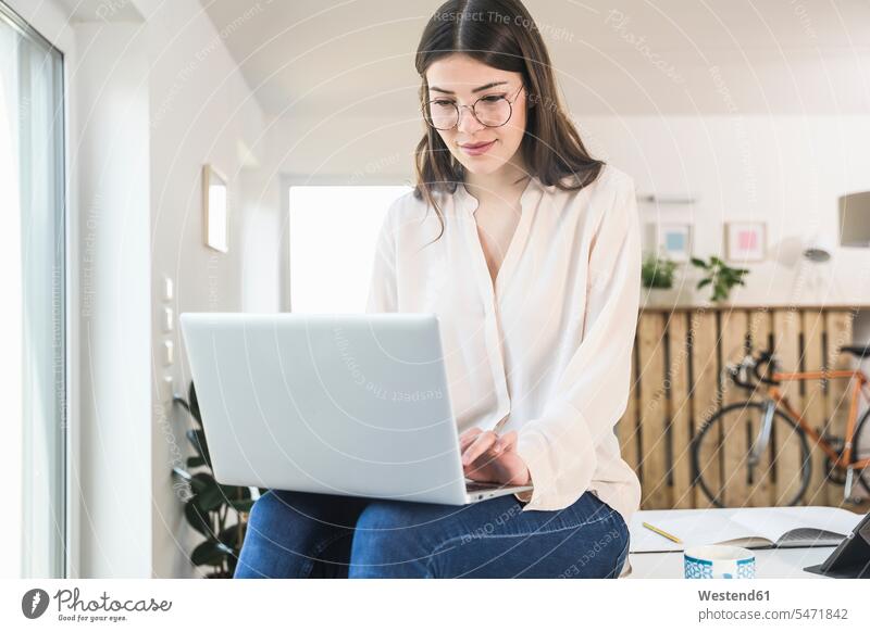 Young woman sitting on table at home using laptop Seated Table Tables Laptop Computers laptops notebook females women computer computers Adults grown-ups