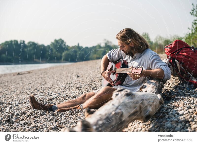 Young man sitting at the riverside playing guitar River Rivers riverbank men males Seated guitars water waters body of water water's edge waterside shore Adults