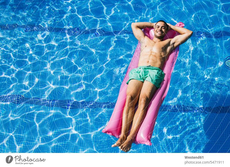 Shirtless handsome young man with hands behind head relaxing on airbed in swimming pool color image colour image Spain day daylight shot daylight shots