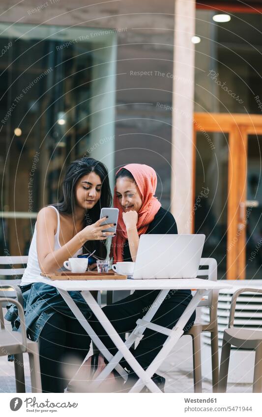 Two friends sitting together at a pavement cafe using cell phone using laptop using a laptop Using Laptops Table Tables Instant Messaging Messenger Seated