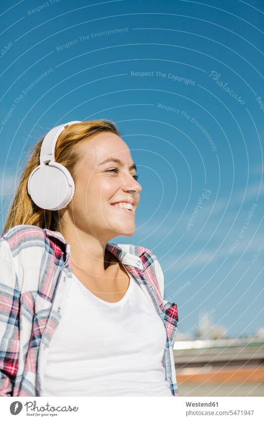 Happy woman listening music through headphone looking away against blue sky on sunny day color image colour image outdoors location shots outdoor shot