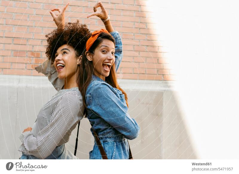 Cheerful friends pointing towards each other while standing back to back against wall color image colour image outdoors location shots outdoor shot