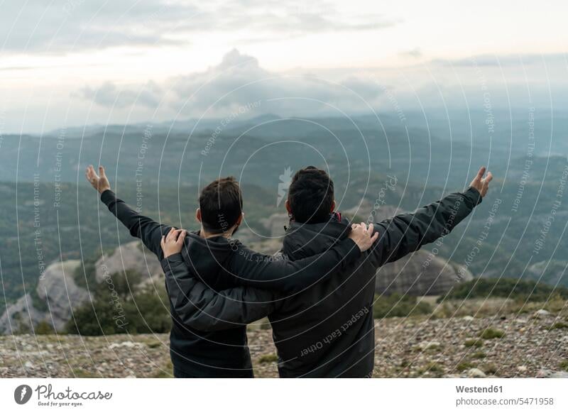 friends standing on mountain peak with arms around, enjoying he view cheering jubilate rejoicing rejoice exultation jubilating casual leisure wear