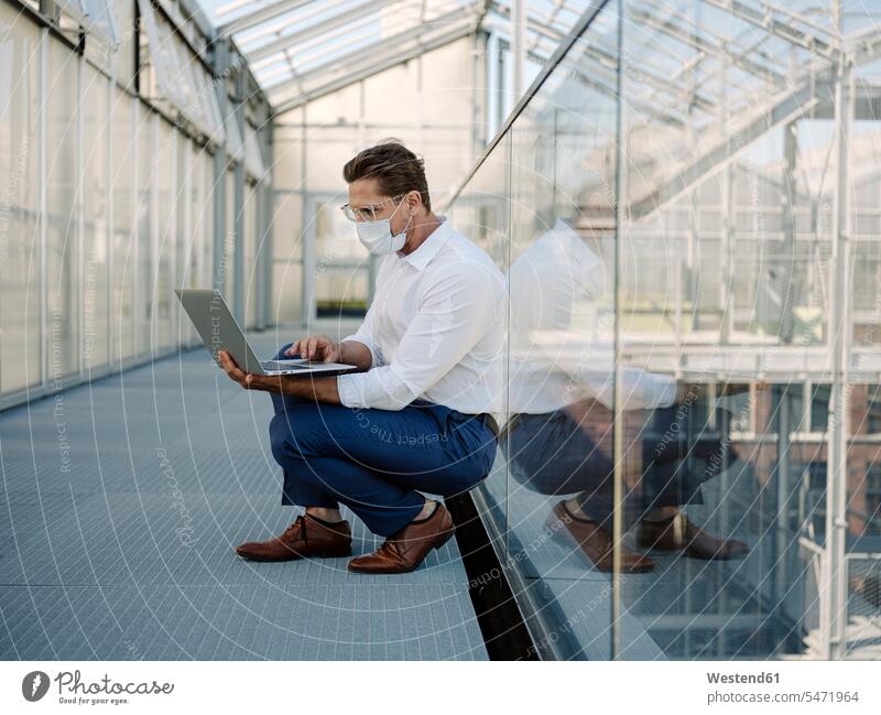 Businessman wearing mask using laptop while crouching on floor in greenhouse color image colour image Germany business people businesspeople