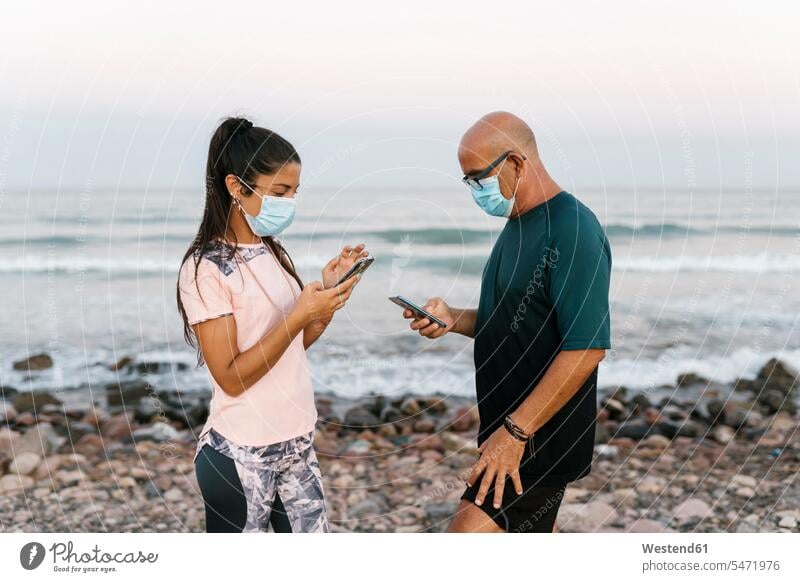 Woman and senior man with face mask using phone while standing against sea color image colour image outdoors location shots outdoor shot outdoor shots sunset