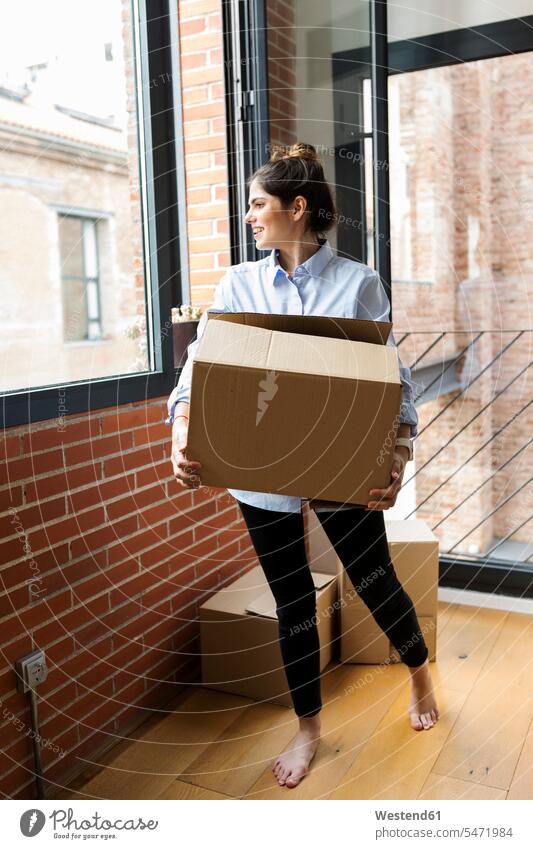 Smiling young woman carrying cardboard box in new apartment looking out of window Cardboard Carton carton cardboard boxes Cardboards cartons females women flats