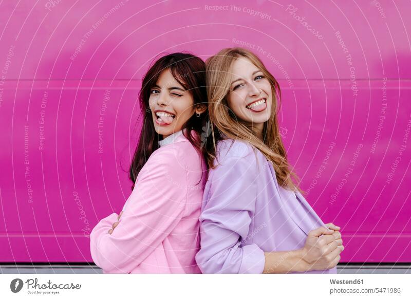 Sisters sticking out tongue while standing back to back by pink wall color image colour image outdoors location shots outdoor shot outdoor shots day