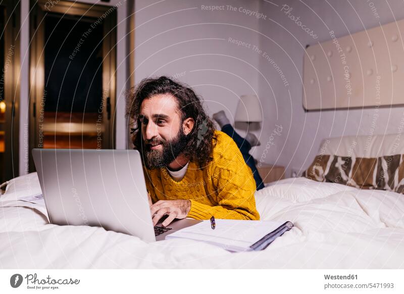 Smiling young man lying in bed at home using laptop beds men males smiling smile Laptop Computers laptops notebook laying down lie lying down Adults grown-ups