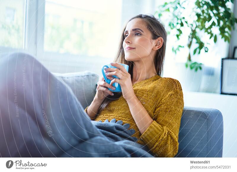 Beautiful young woman enjoying cup of coffee while lying on sofa under blanket human human being human beings humans person persons celibate celibates singles