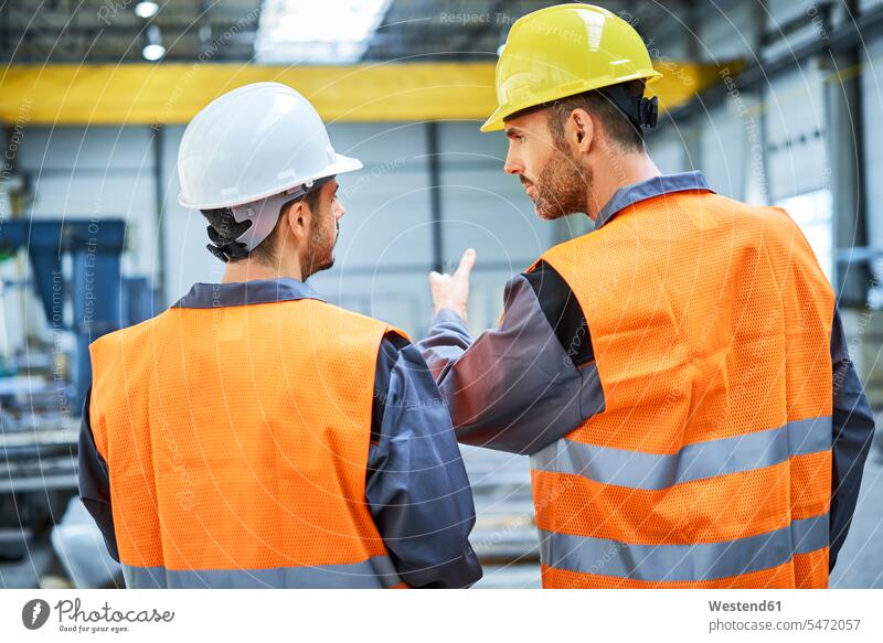 Rear view of two men wearing protective workwear talking in factory Protective Workwear Protective Work Wear man males speaking industry industrial Adults