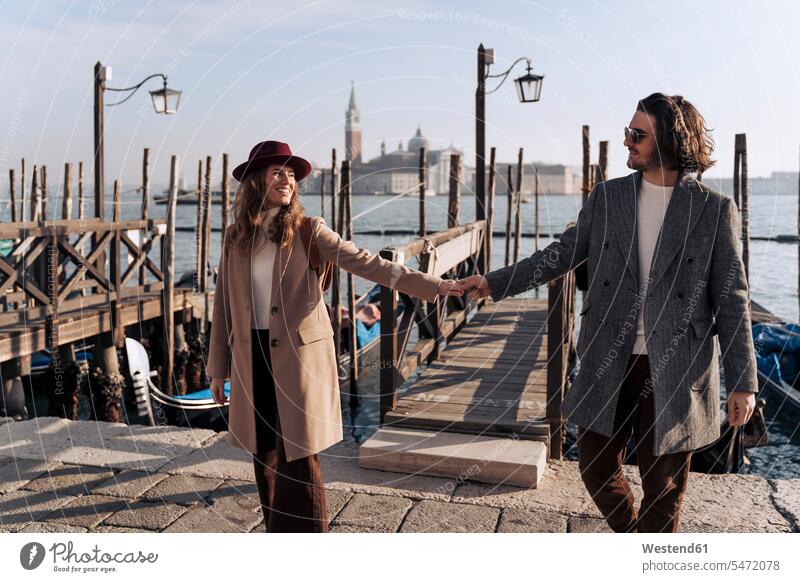 Young couple walking hand in hand at the waterfront in Venice, Italy hats coat coats jackets Eye Glasses Eyeglasses specs spectacles Pair Of Sunglasses