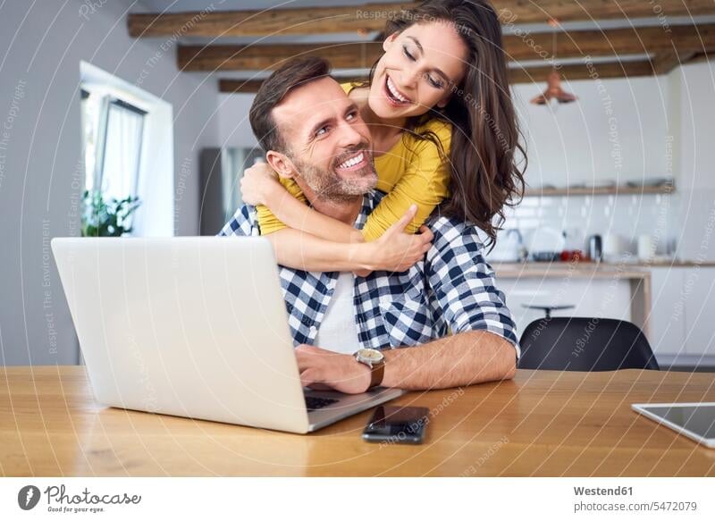 Happy couple sitting at dining table, embracing, using laptop using a laptop Using Laptops happiness happy Table Tables Laptop Computers laptops notebook