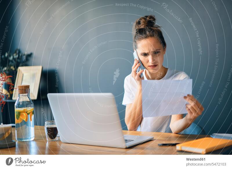 Businesswoman holding document talking over smart phone while sitting at desk in home office color image colour image casual clothing casual wear leisure wear