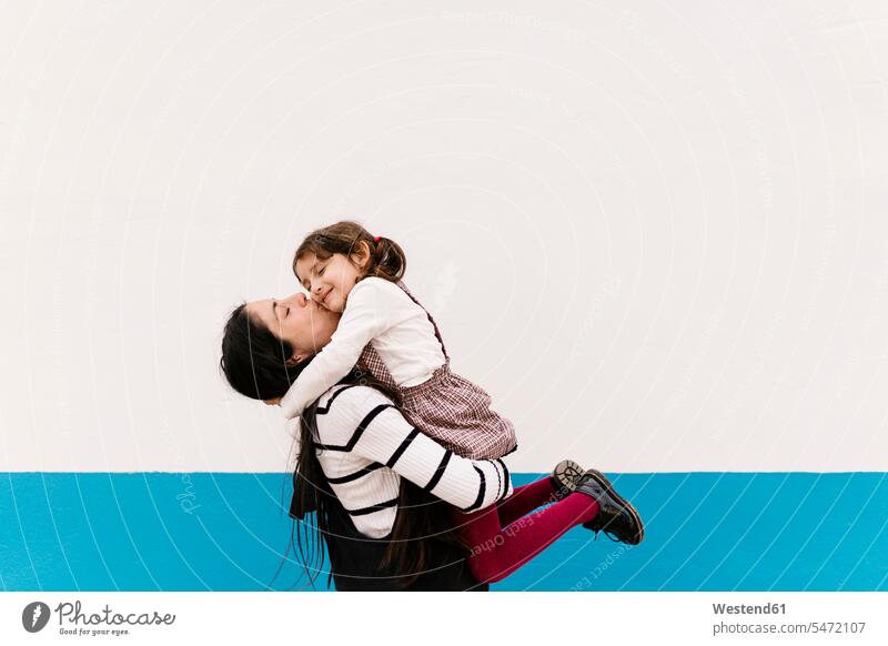 Mother kissing daughter on cheek against wall during sunny day color image colour image outdoors location shots outdoor shot outdoor shots daylight shot