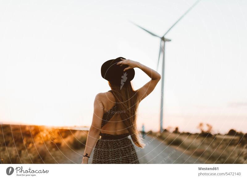 Back view of young woman on rural road in the evening with wind wheel in the background country road rural roads country roads windmills wind turbine