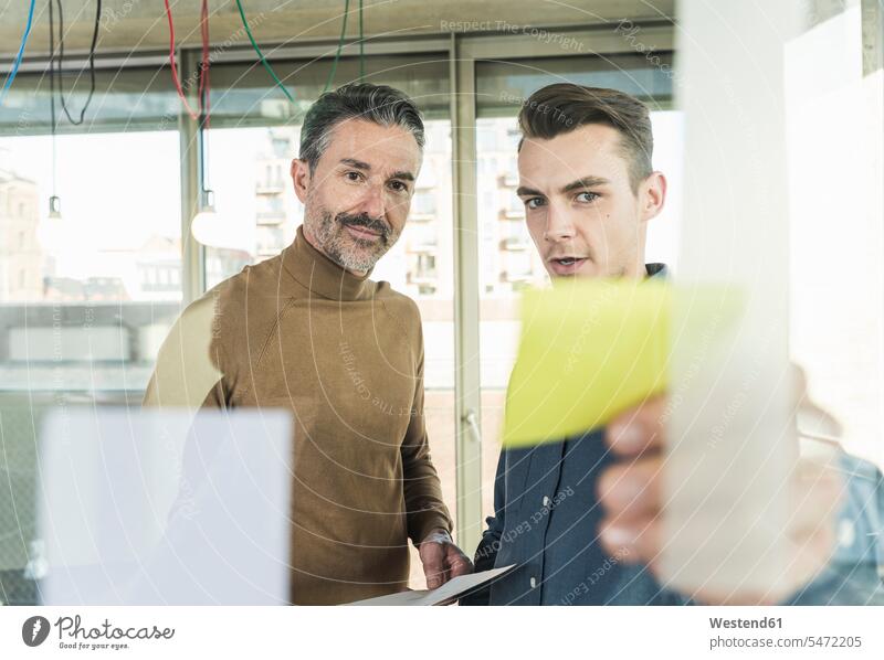 Mature businessman and young man working on adhesive notes at glass pane in office colleague associate associates partner partners partnerships Occupation Work