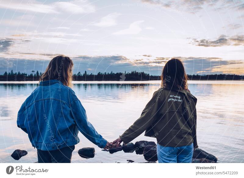 Girl friends looking at Lake Inari,Finland, holding hands lake lakes sitting Seated young women young woman hand in hand water waters body of water females