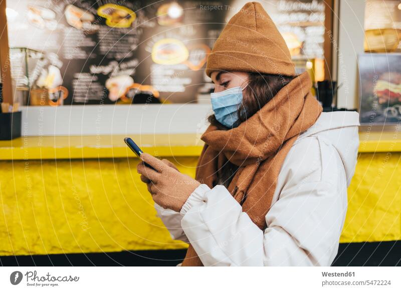 Young woman using smart phone waiting outside cafe in winter during COVID-19 color image colour image outdoors location shots outdoor shot outdoor shots day