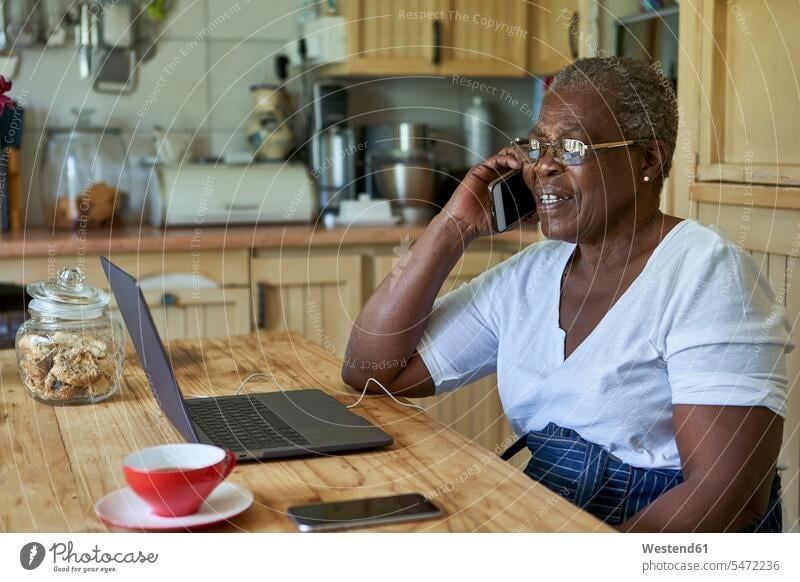 Senior woman sitting at kitchen table using laptop and smartphone human human being human beings humans person persons African black black ethnicity coloured 1