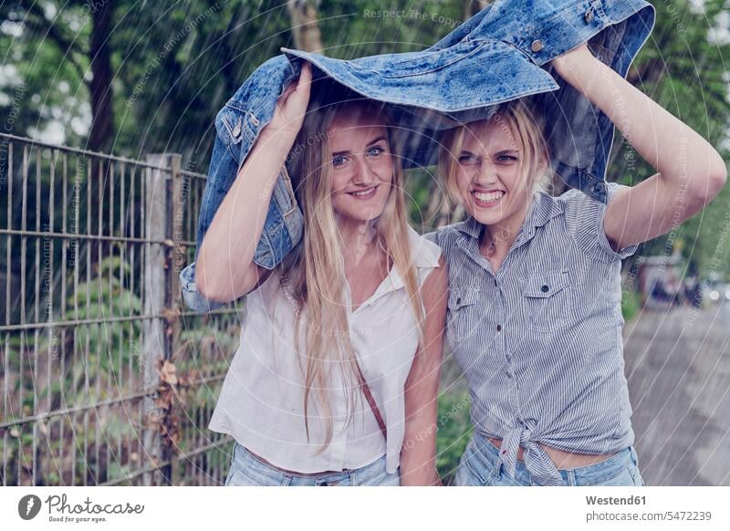 Two young women shletering from rain with a denim jacket female friends jeans jackets woman females hedge hedges Hedgerow protecting guarding shelter sheltering