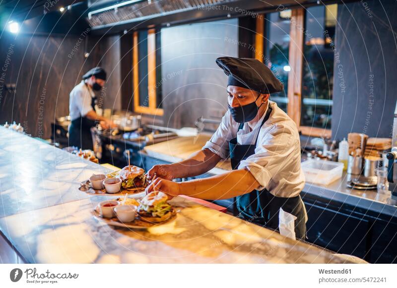 Chef wearing protective face mask preparing burgers in restaurant kitchen human human being human beings humans person persons 2 2 people 2 persons two