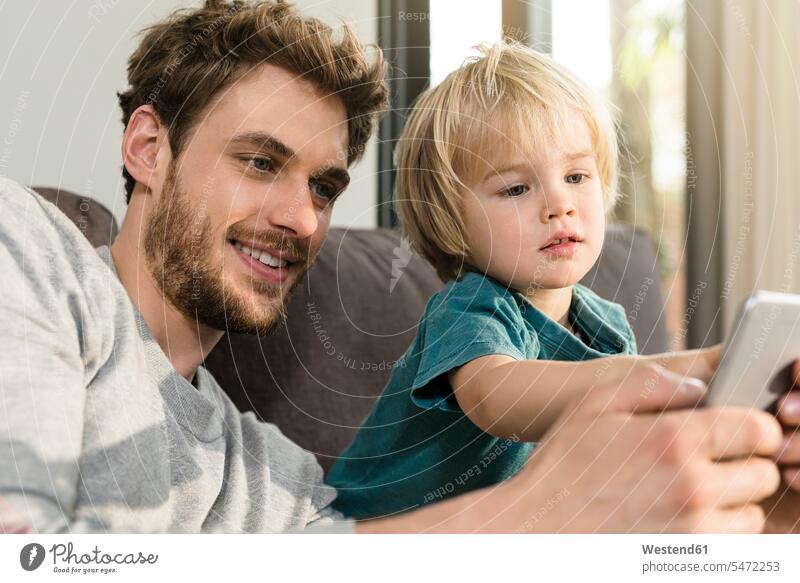 Father and son looking at smartphone on couch at home settee sofa sofas couches settees eyeing father pa fathers daddy dads papa Smartphone iPhone Smartphones