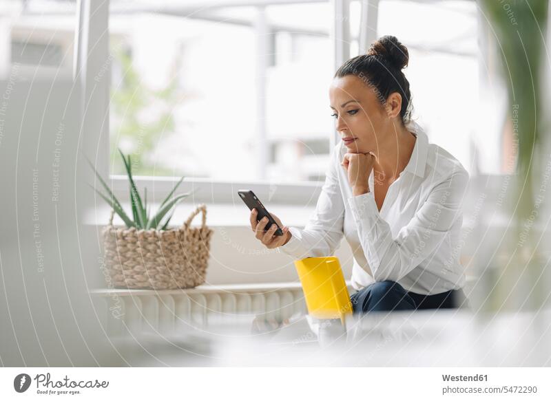 Female entrepreneur using mobile phone while sitting by window in home office color image colour image Germany indoors indoor shot indoor shots interior