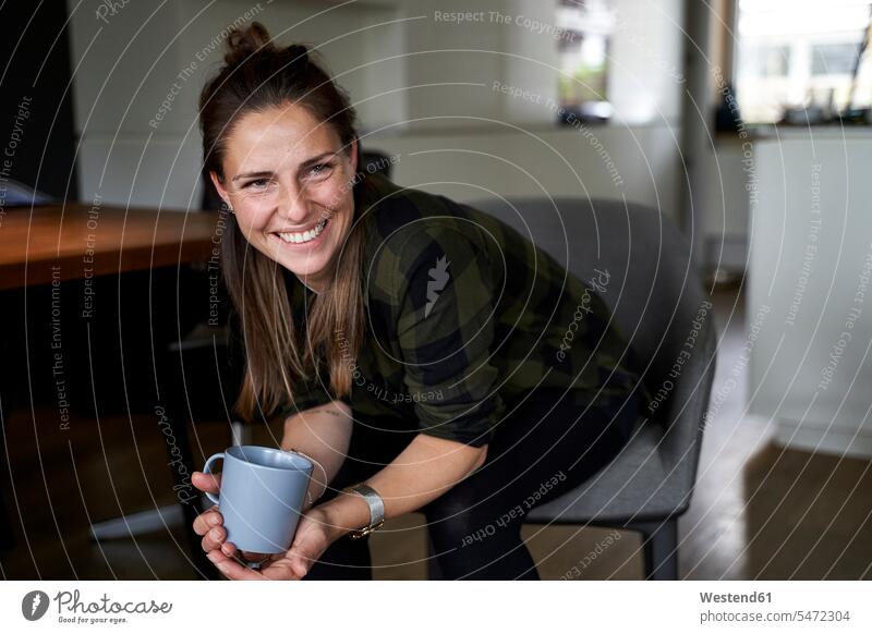 Smiling woman holding coffee mug looking away while sitting on chair at home color image colour image Germany leisure activity leisure activities free time