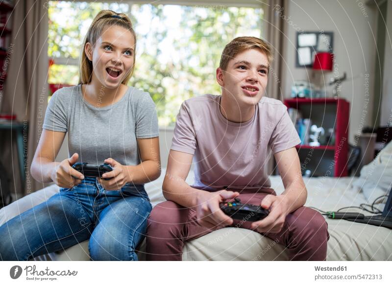 Happy teenage girl and boy sitting on bed playing video game beds friends mate video games Seated watching happiness happy boys males Teenage Girls