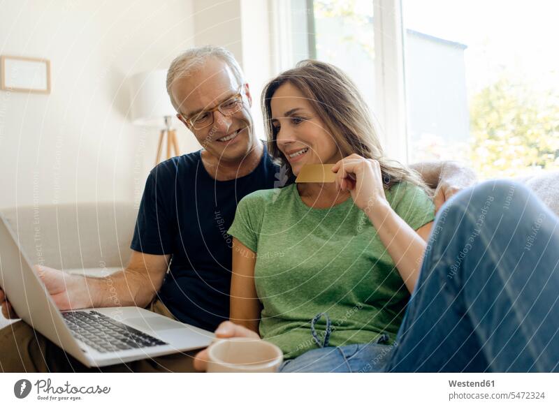 Smiling mature couple sitting on couch at home shopping online with laptop couches settee settees sofa sofas computers Laptop Computer Laptop Computers laptops