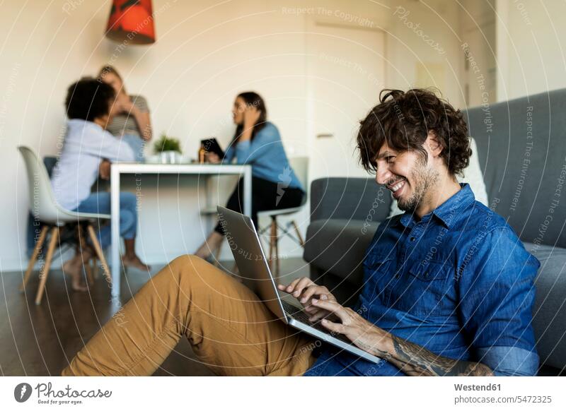 Smiling man sitting on floor using laptop with friends in background floors Laptop Computers laptops notebook men males Seated smiling smile friendship computer