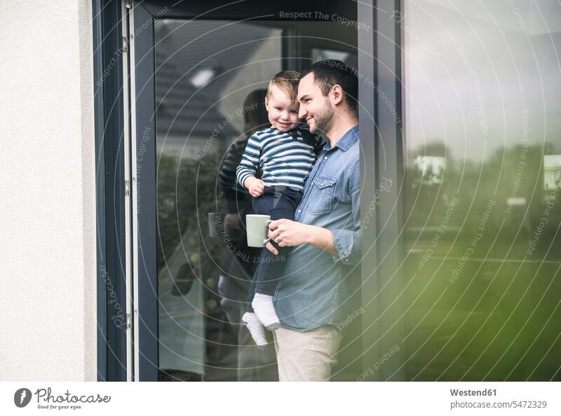 Father with cup of coffee carrying son at terrace door at home patio door French window father fathers daddy dads papa sons manchild manchildren Coffee Cup