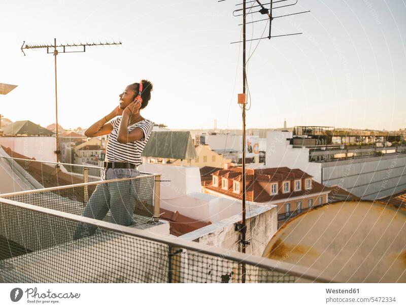 Happy young woman standing on rooftop at sunset listening to music with headphones human human being human beings humans person persons African black