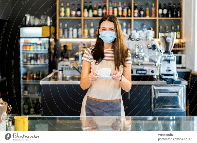 Beautiful owner wearing mask while holding coffee cup and saucer in cafe color image colour image indoors indoor shot indoor shots interior interior view