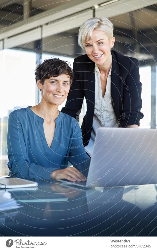 Portrait of two smiling businesswomen with laptop at desk in office smile offices office room office rooms businesswoman business woman business women