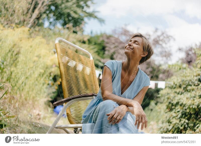 Relaxed woman sitting in garden on chair with closed eyes relaxed relaxation Seated chairs females women gardens domestic garden relaxing Adults grown-ups