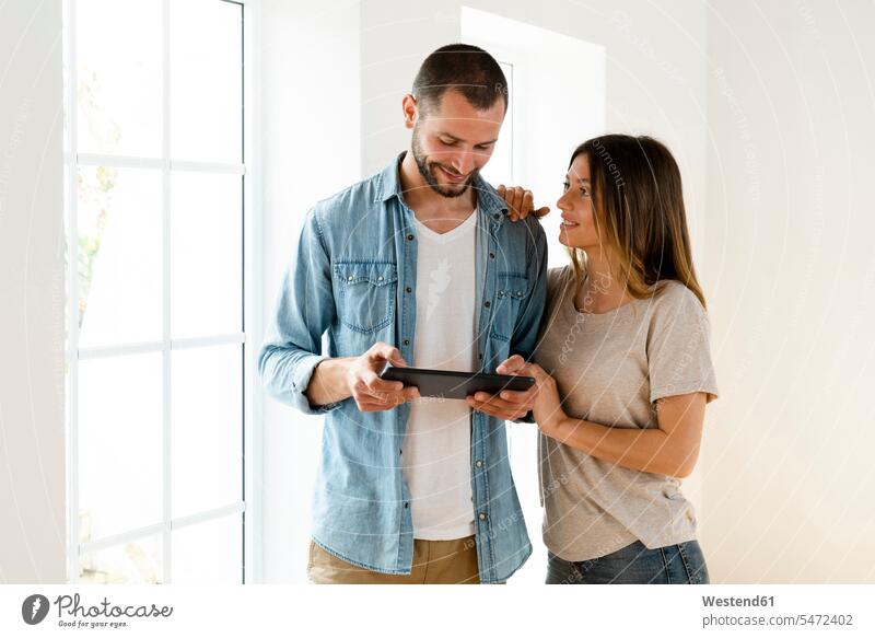 Smiling couple at home standing in front of window looking at tablet together windows hold smile speak speaking talk relax relaxing relaxation delight enjoyment
