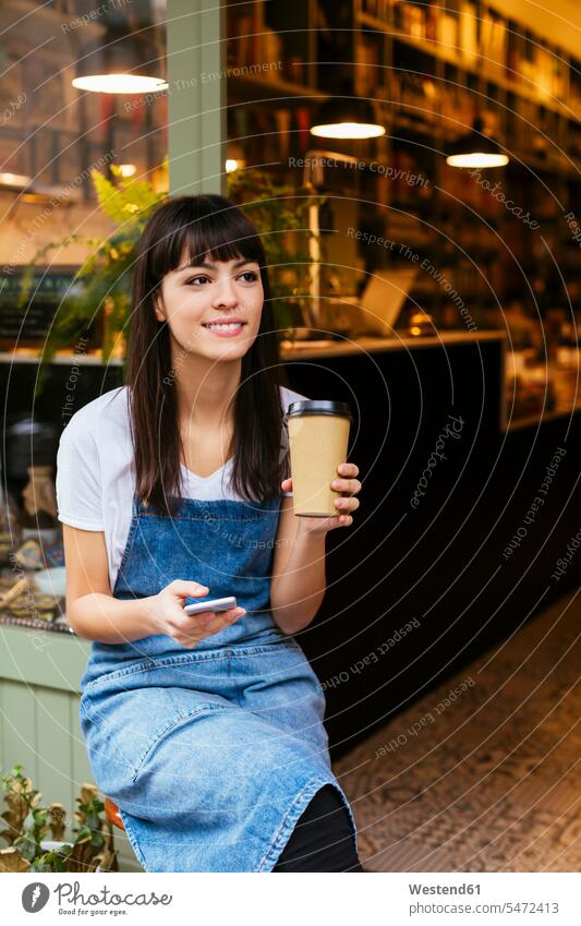Smiling woman sitting at entrance door of a store holding cell phone and takeaway coffee Coffee Seated smiling smile shop females women mobile phone mobiles