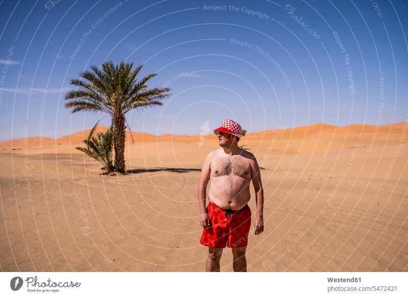 Overweight man with swimming shorts and hat standing in the desert of Morocco (value=0) touristic tourists Helplessness swim wear bathing trunks swim trunks
