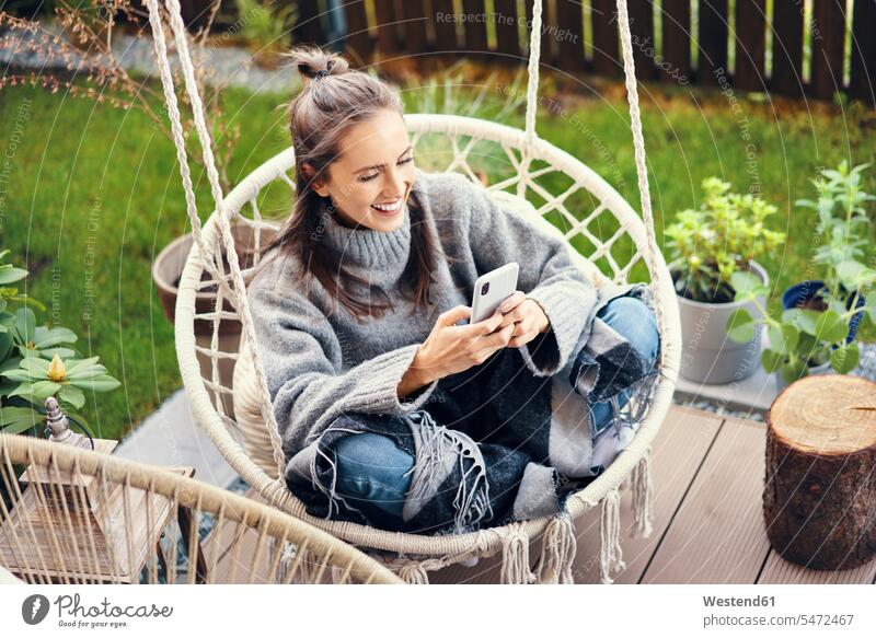 Happy young woman using smart phone while sitting on swing in garden caucasian caucasian appearance caucasian ethnicity european White - Caucasian caucasians