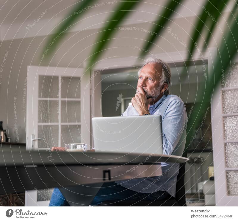 Pensive mature man sitting at table with laptop in his living room pensive thoughtful Reflective contemplative Laptop Computers laptops notebook men males Table