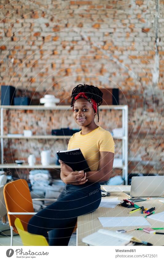 Portrait of confident young woman at table in office holding notebook human human being human beings humans person persons African black black ethnicity