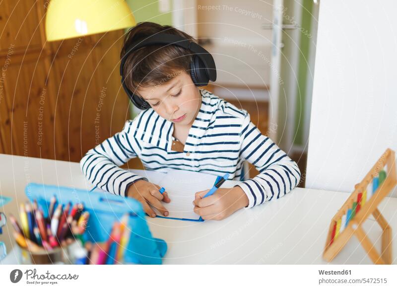 Boy doing homeschooling and writing on notebook, using tablet and headphones at home Tables desks pencil pencils pens headset hear learn write free time