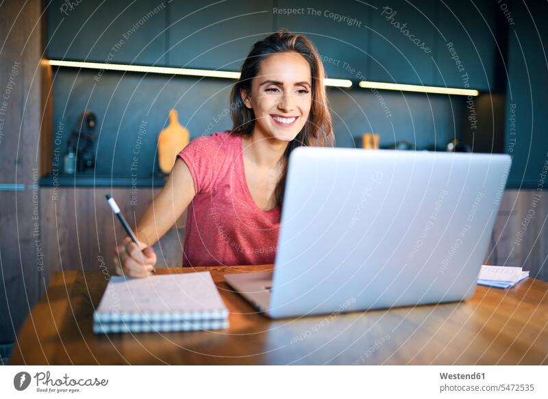 Smiling female entrepreneur looking at laptop while writing in note pad on desk color image colour image casual clothing casual wear leisure wear casual clothes