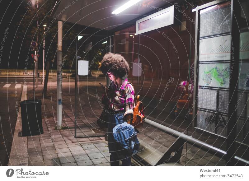 Young woman with afro hairdo using smartphone at bus stop in the city human human being human beings humans person persons curl curled curls curly hair